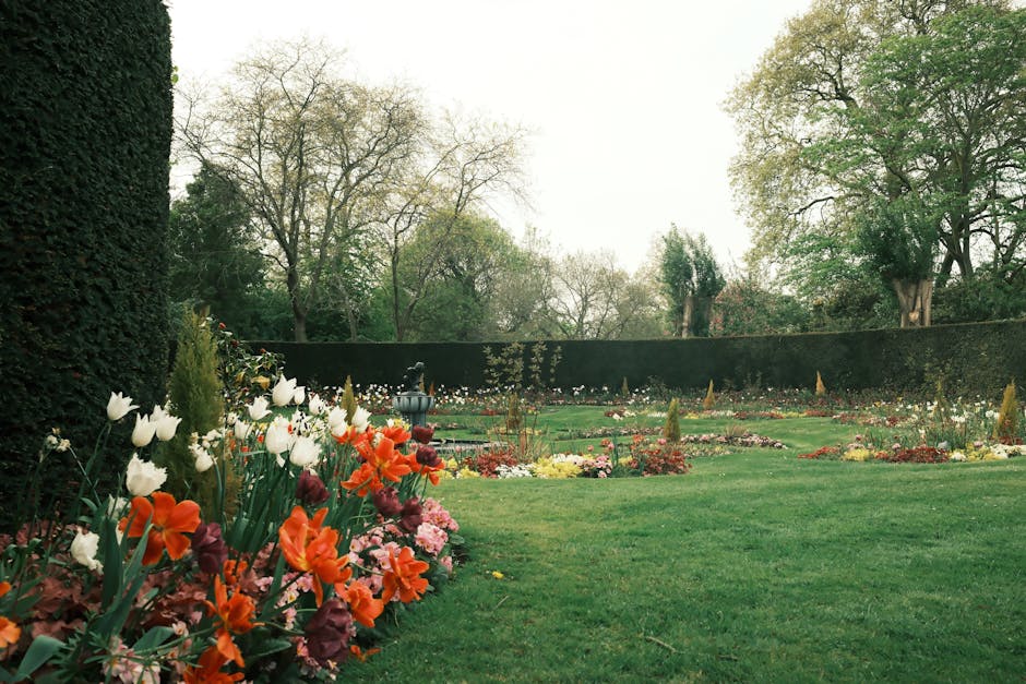 A well-maintained garden scene with lush, trimmed green grass covering the foreground, bordered by colorful flower beds containing vibrant tulips, primroses, and other seasonal blooms. To the left, a 